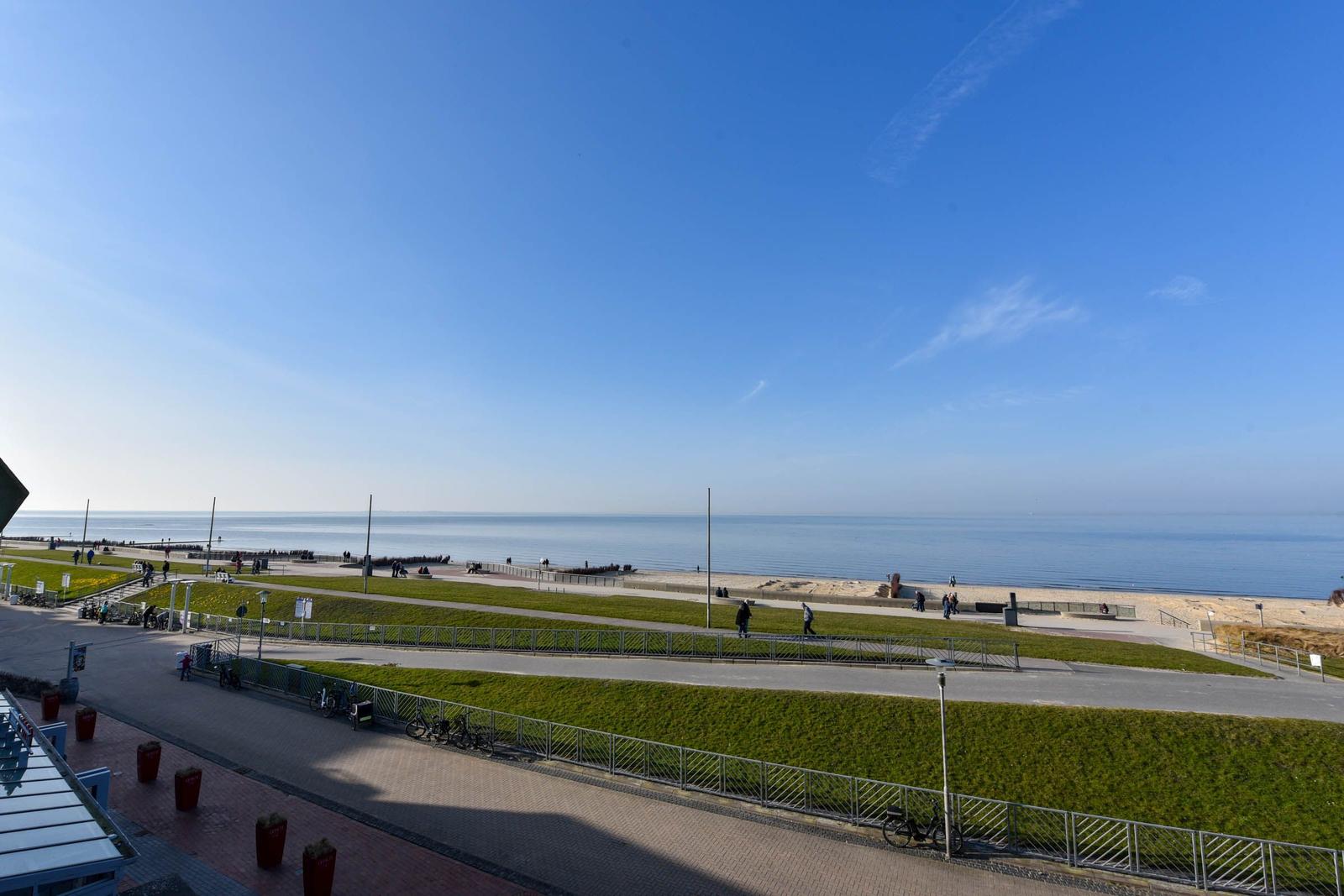 Meerfront mit Strand, Grünflächen und Spaziergängern unter blauem Himmel.