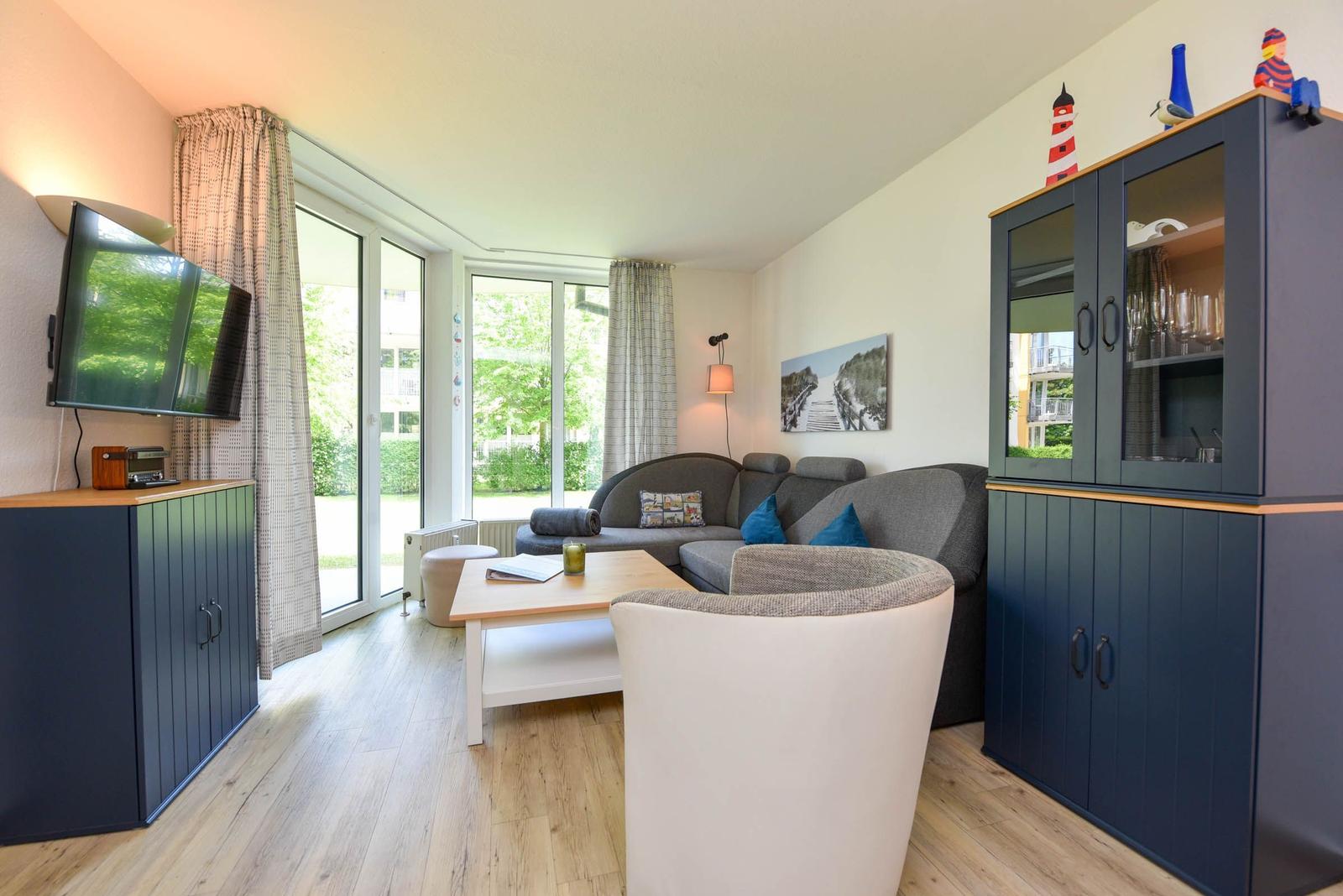 Wohnzimmer mit Sofa, Sessel, Couchtisch und Glaswand-Schrank. Fenster mit Blick auf Garten.