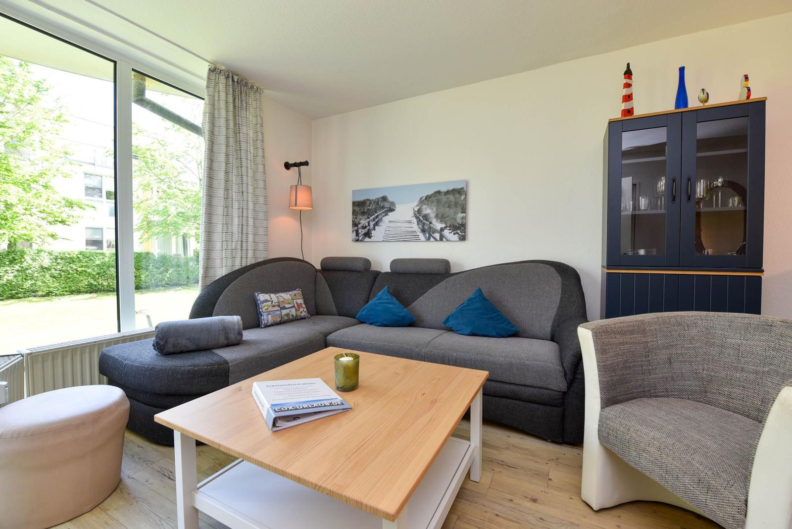 Wohnzimmer mit grauem Sofa, Holztisch und Glaswand-Schrank. Große Fenster mit Blick auf Grün.