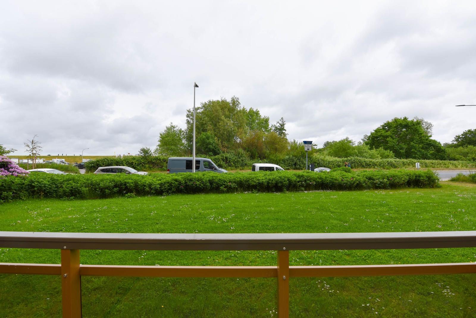 Grünes Feld mit Hecke und Parkplatz im Hintergrund. Gelber Geländer im Vordergrund.