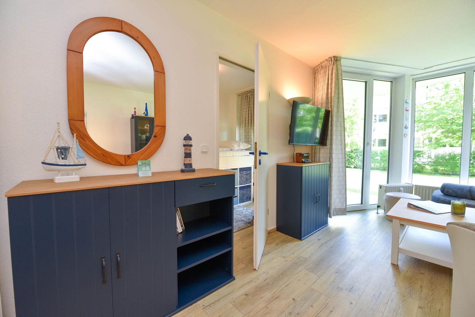 Wohnzimmer mit Fernseher, Schrank und Blick auf Garten. Tür führt in Schlafzimmer.
