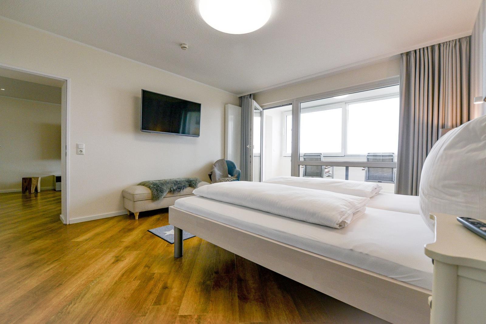 Bedroom with double bed, TV, and balcony view. Wooden floor and light walls.