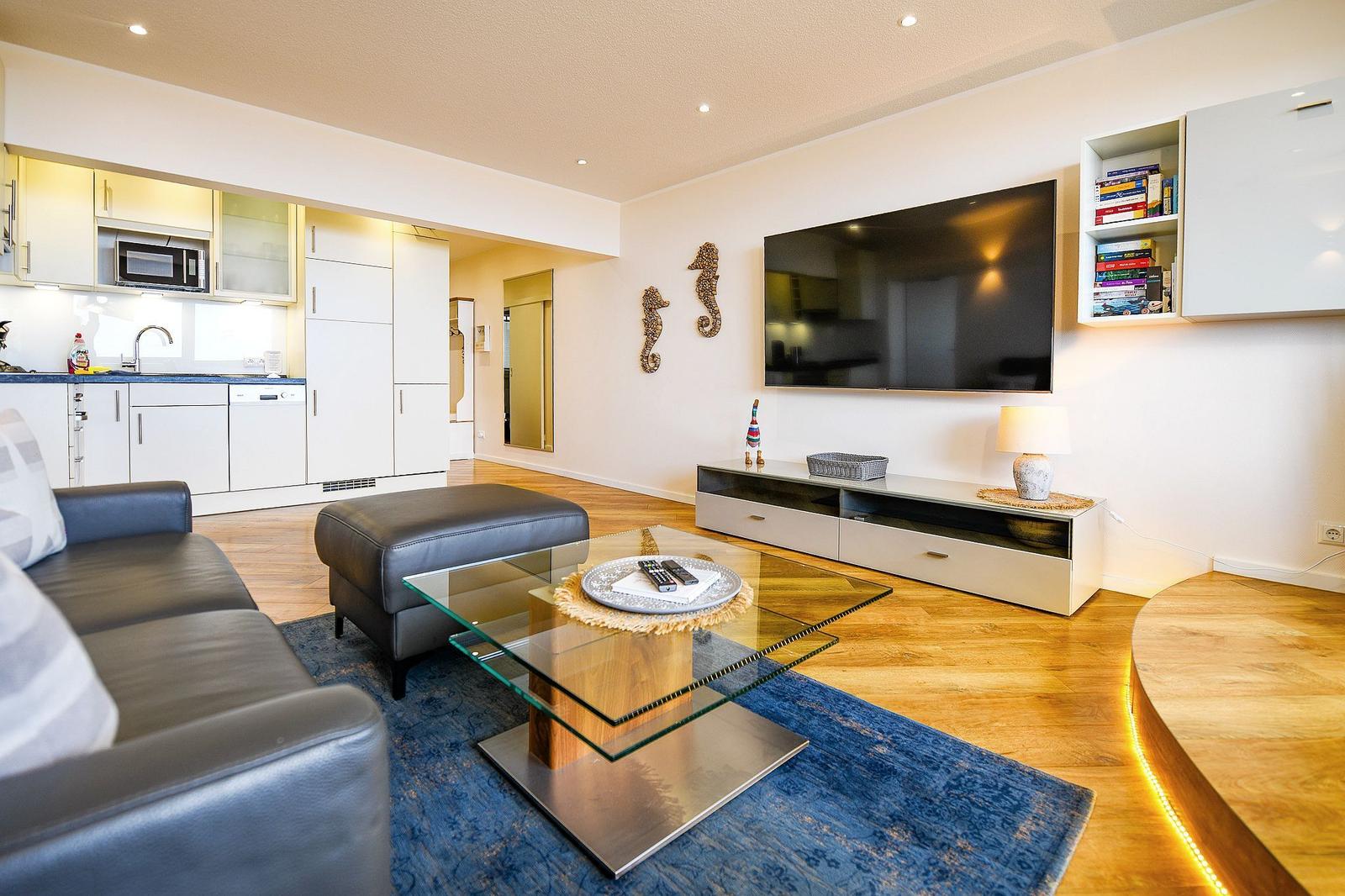 Living room with sofa, glass table, and TV. Kitchen in background. Wooden floor and rug.