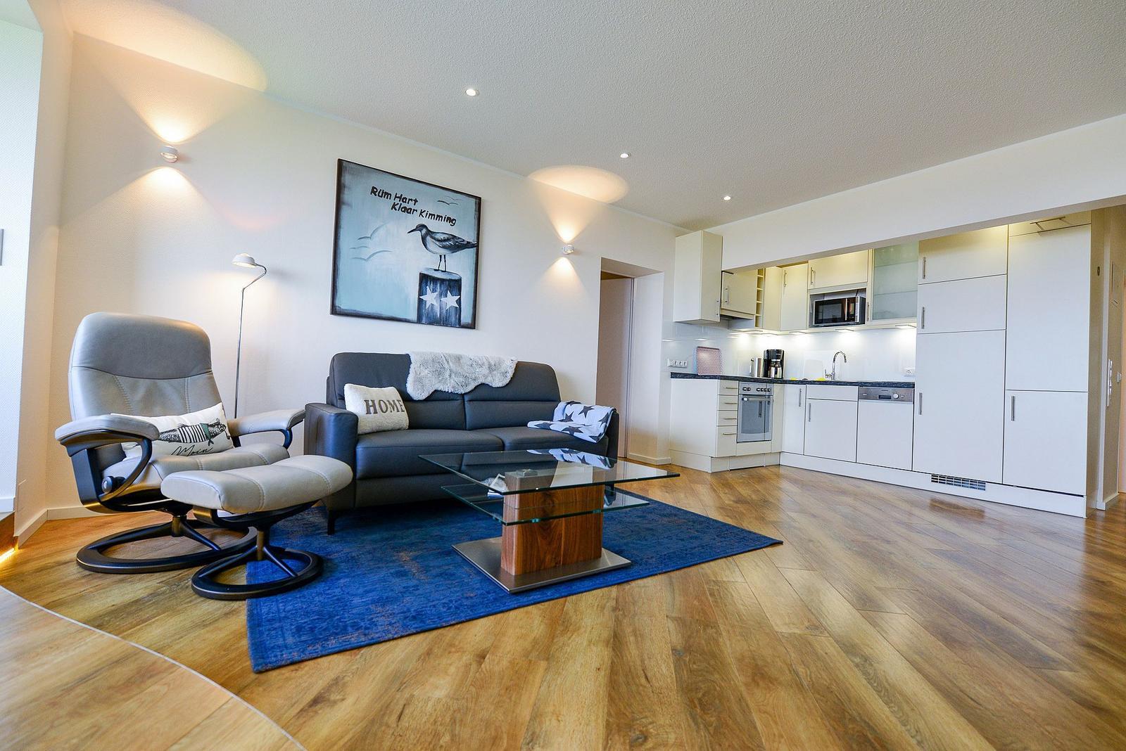 Living room with sofa, armchair, and glass table on blue rug. Kitchen with white cabinets in background.