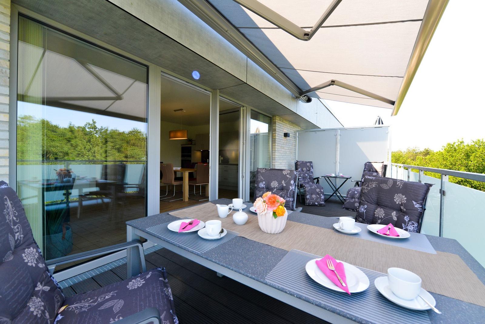 Terrasse mit Tisch, Stühlen und Sonnenschutz. Blick ins Haus und auf grüne Umgebung.