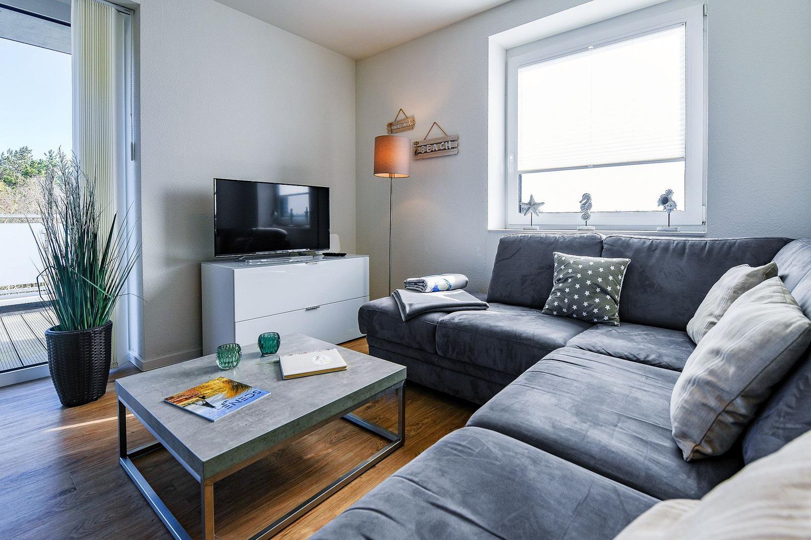 Ein Wohnzimmer mit grauem Sofa, Couchtisch und Fernseher. Fenster und Balkontür sind sichtbar.
