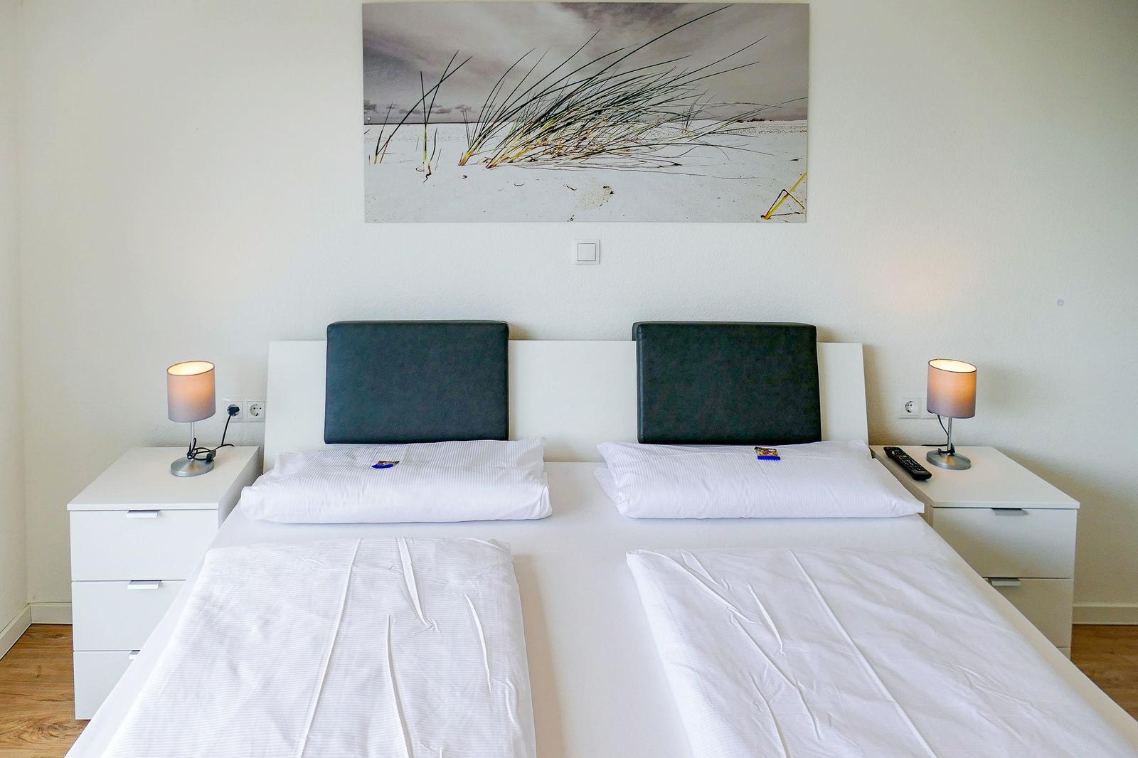 Zwei Betten mit weißen Bettwäsche, Nachttische und Wandbild mit Strandmotiv.