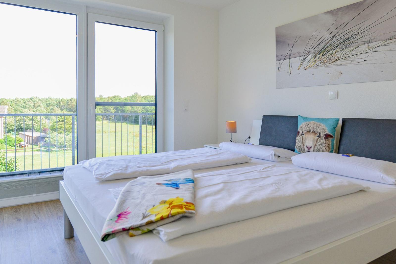Schlafzimmer mit Balkon, Blick auf Wiese und Wald, Bett mit Kissen und Decke.