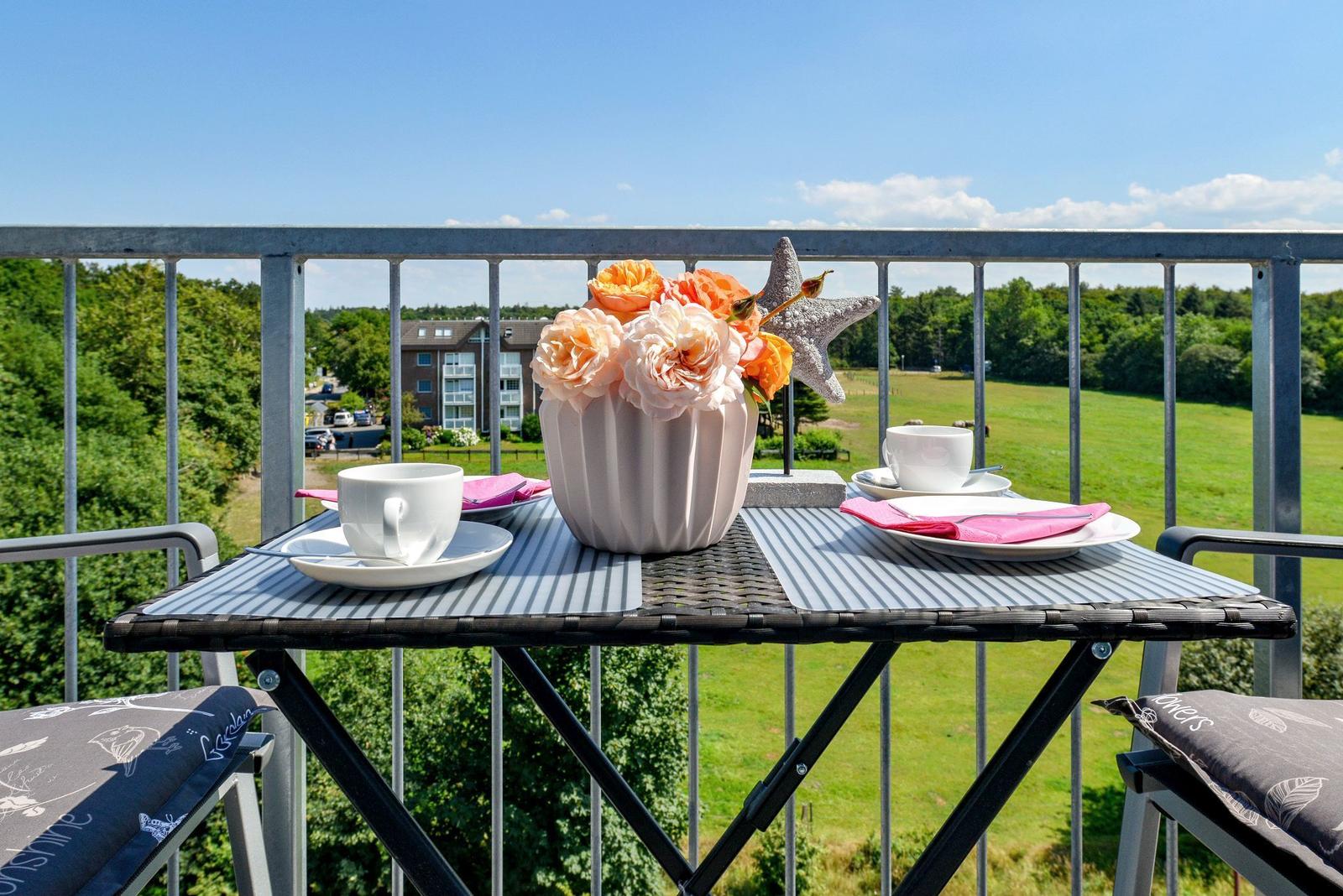 Balcony table set for two with flowers and cups overlooking green fields.