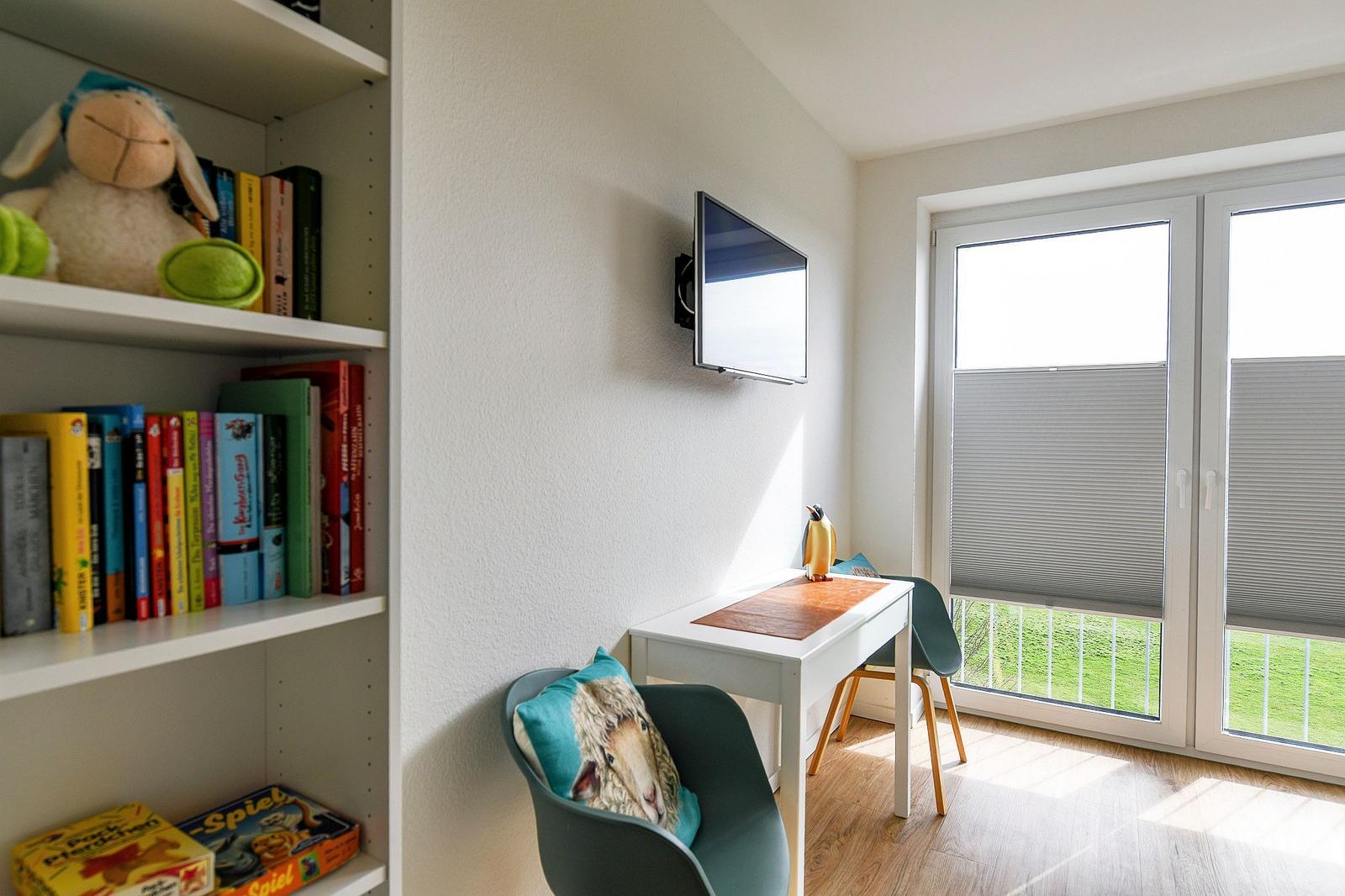 Ein Raum mit Schreibtisch, Fernseher und Bücherregal. Durchgangstür mit Jalousie zum Garten.