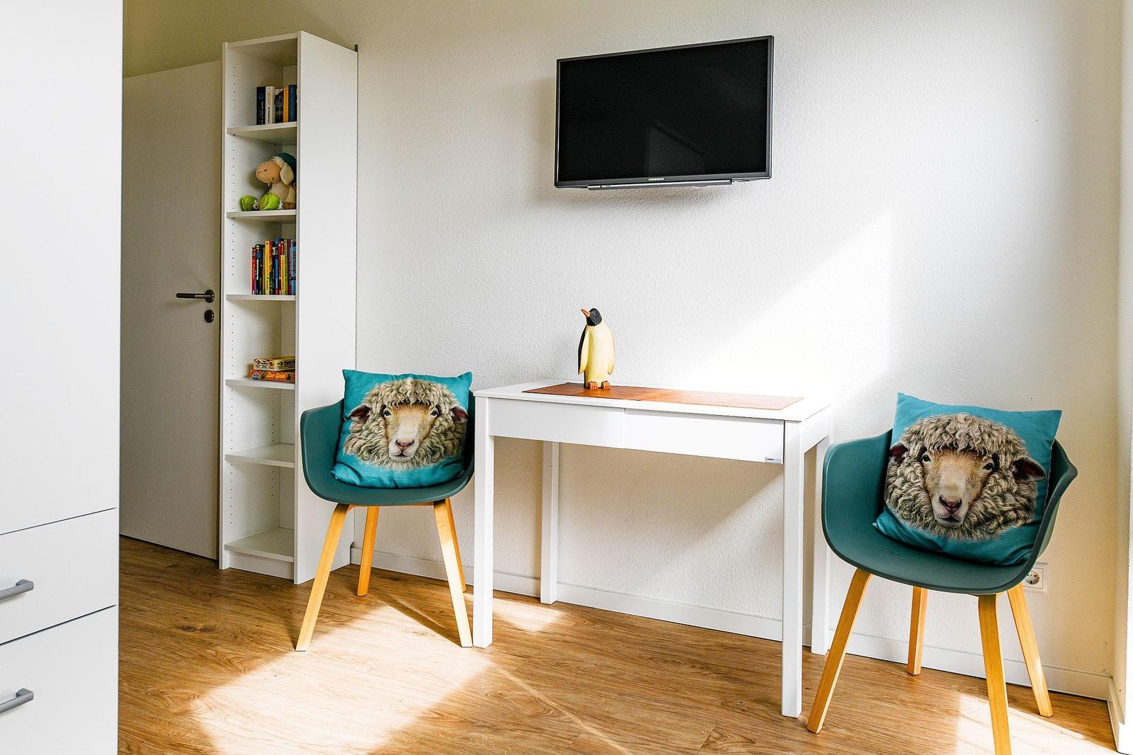 Zwei Stühle mit Schafkissen stehen an einem Schreibtisch unter einer Wand-TV.