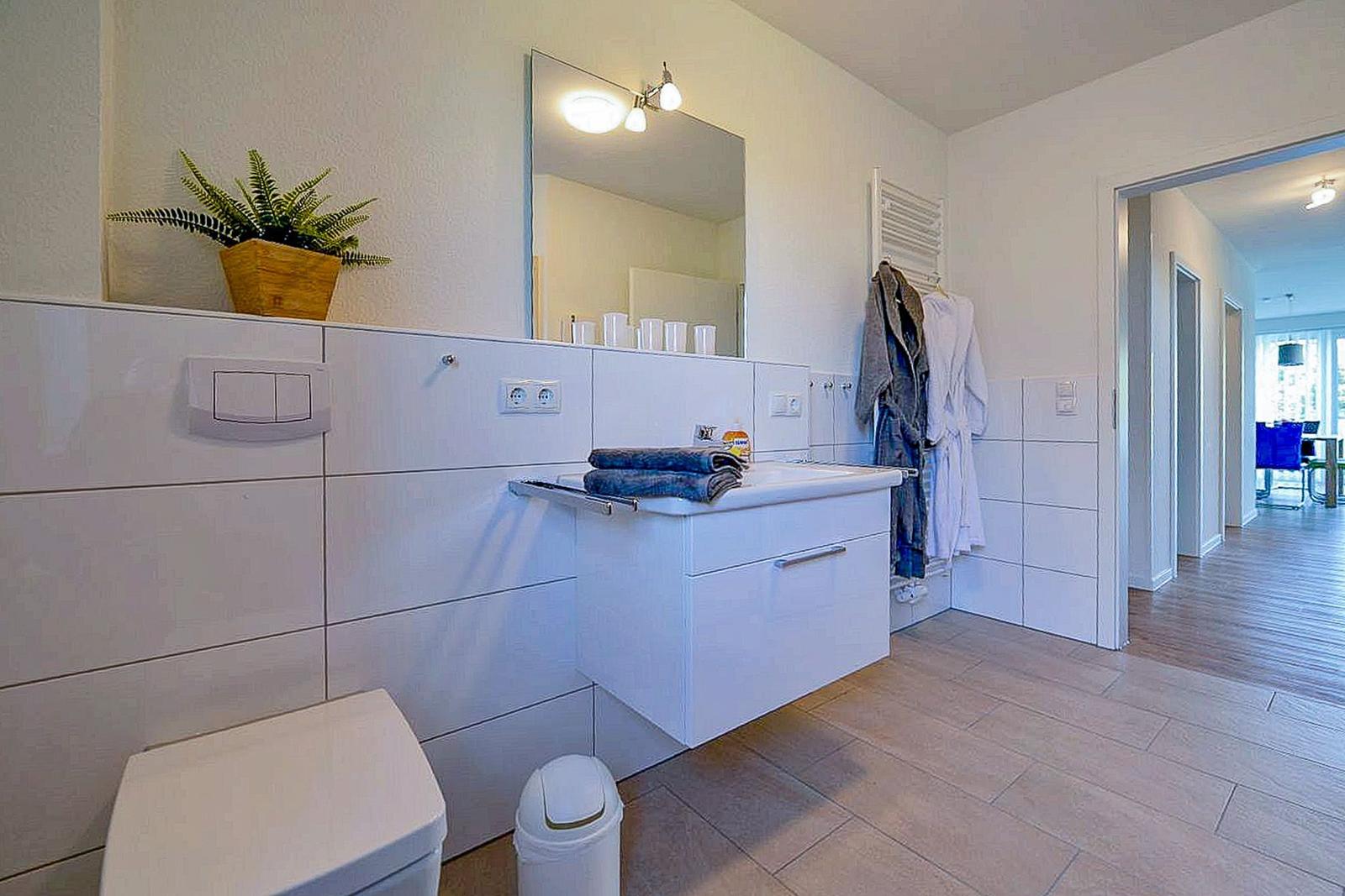 Badezimmer mit Waschbecken, Spiegel, Toilette und Pflanze. Handtücher und Bademäntel hängen bereit.