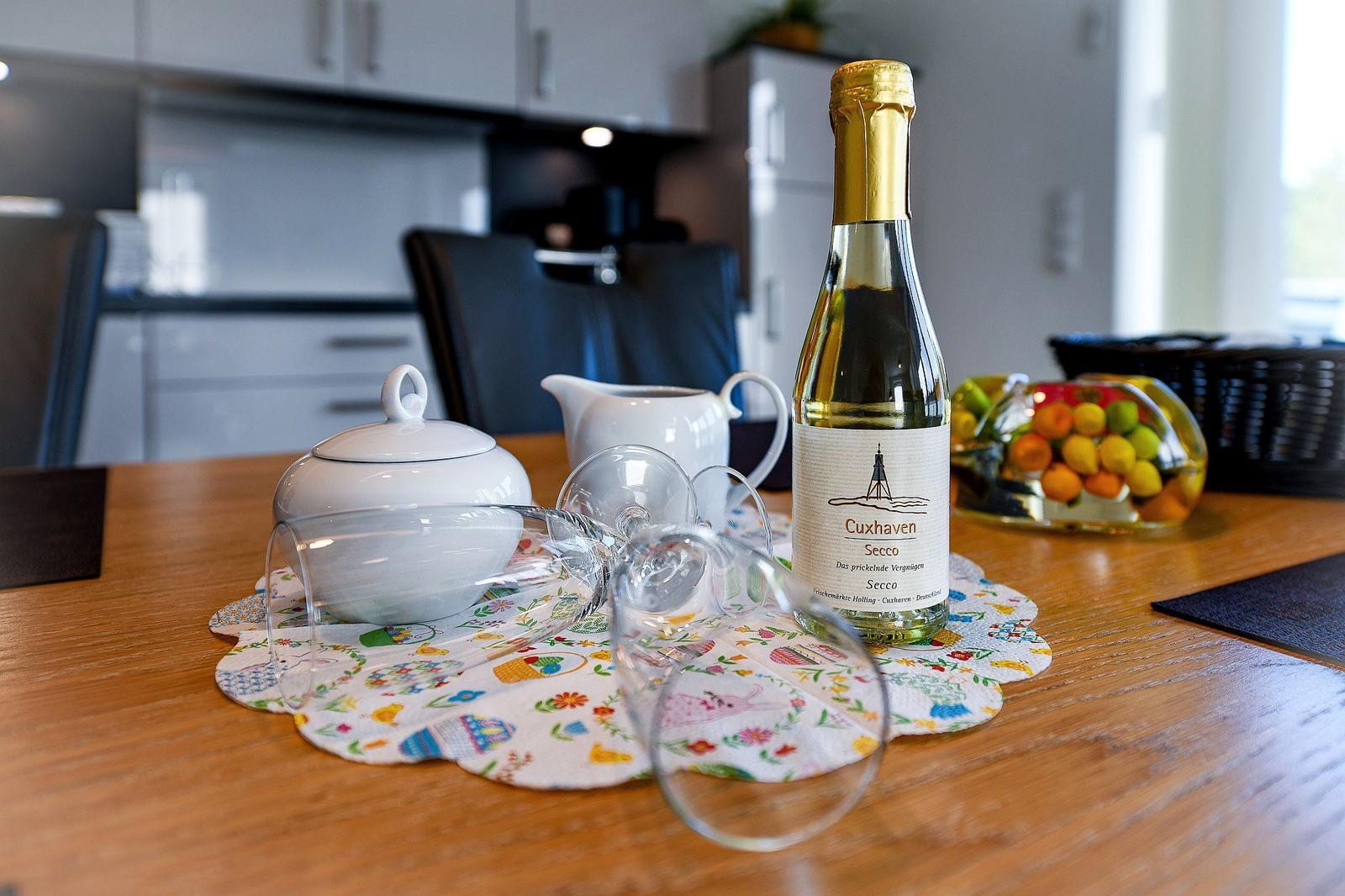 Auf dem Tisch steht eine Flasche Cuxhaven Secco, Gläser, eine Tasse und eine Schale mit Früchten.