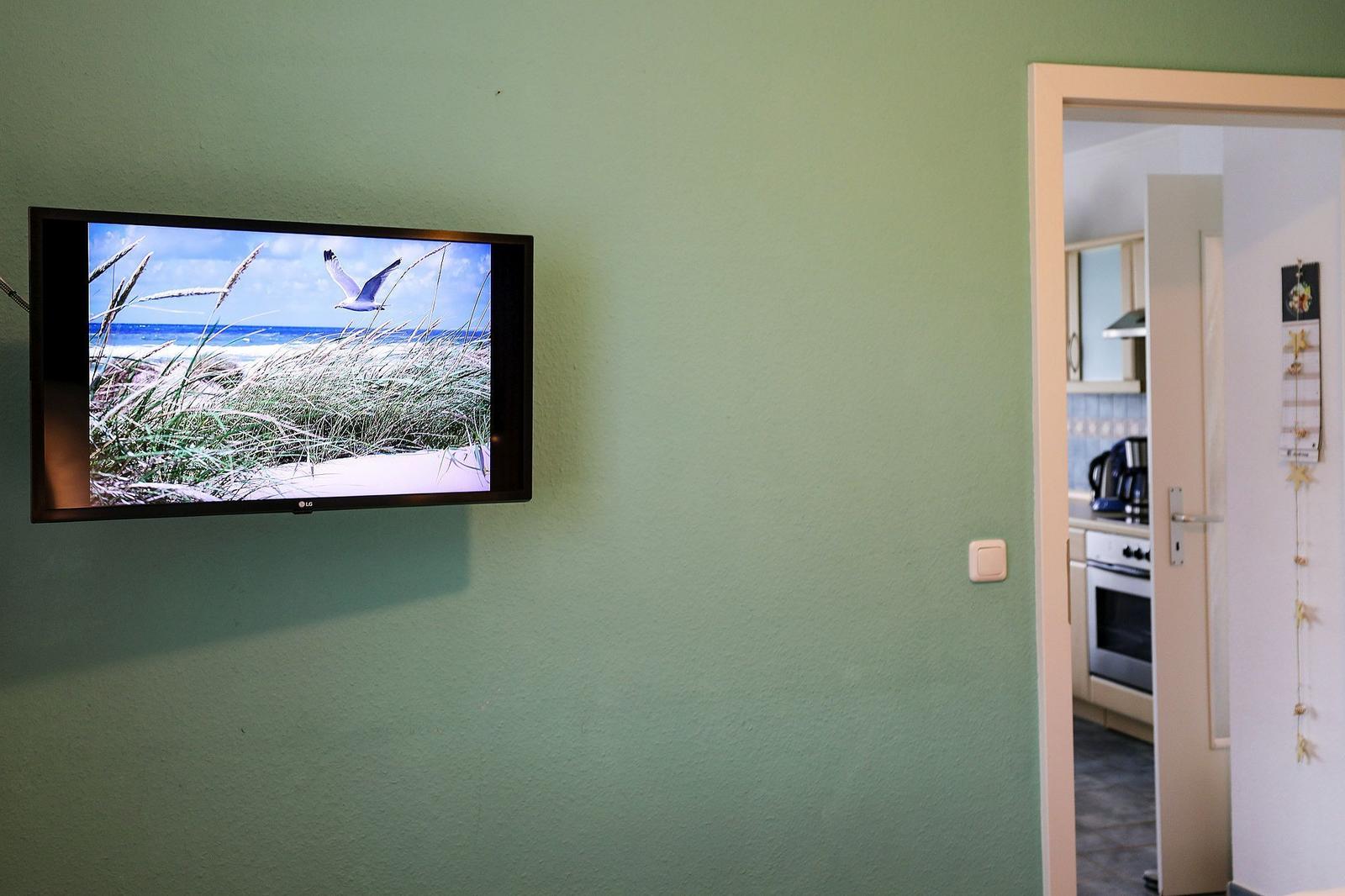 TV an der Wand zeigt Strandbild. Durch Tür ist Küche sichtbar.