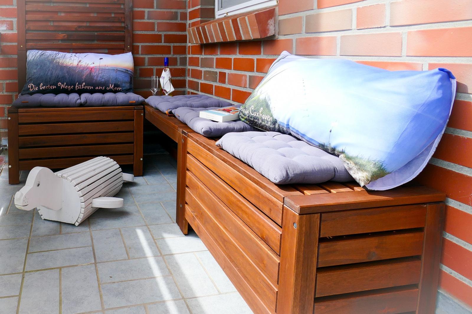 Terrasse mit Holzbank, Kissen und Flasche. Weiße Holzfigur auf Fliesenboden.