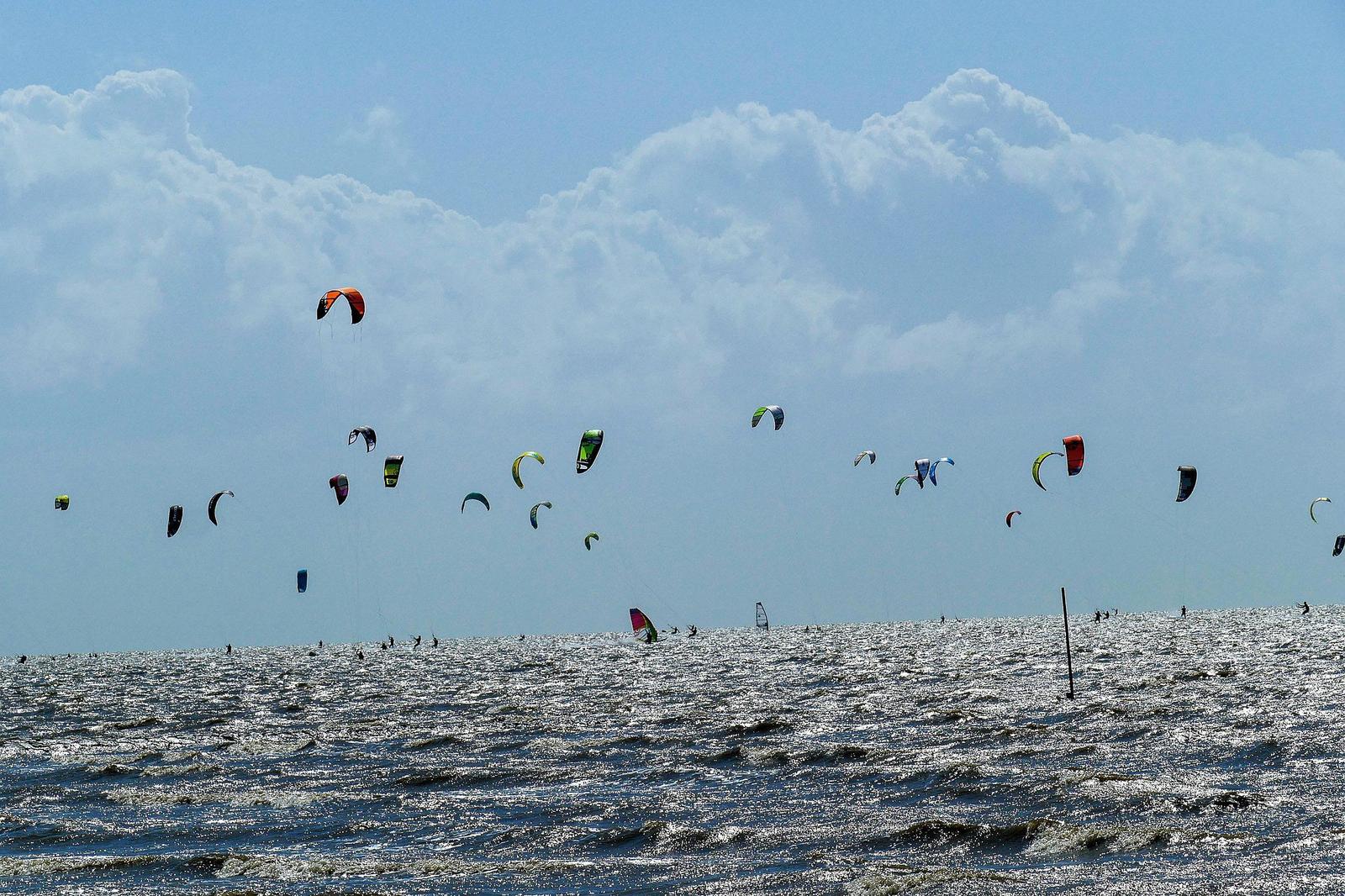 Many kitesurfing kites fly over the sea under a blue sky with white clouds.
