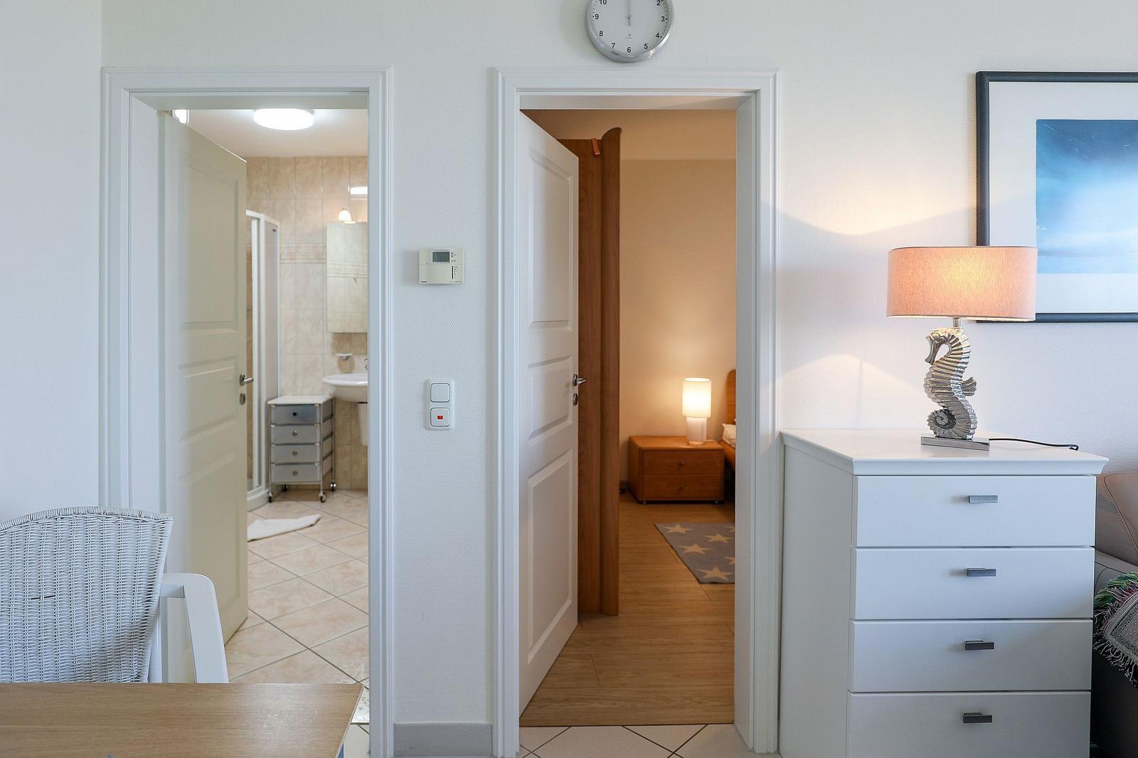 View into bedroom and bathroom. Lamp, dresser, and clock visible.