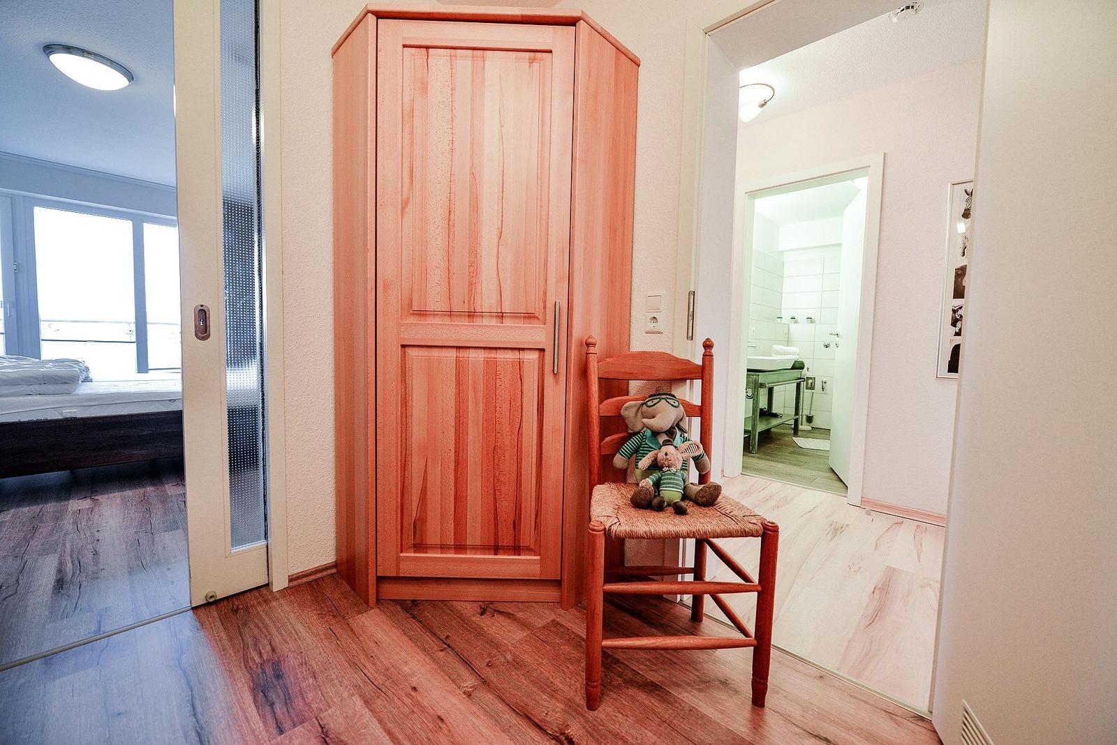 Holzschrank und Stuhl mit Plüschtier im Flur. Blick in Schlafzimmer und Bad.