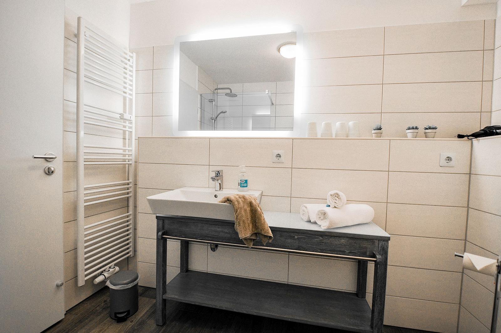 Badezimmer mit Waschbecken, Spiegel und Dusche. Auf der Arbeitsfläche liegen Handtücher.