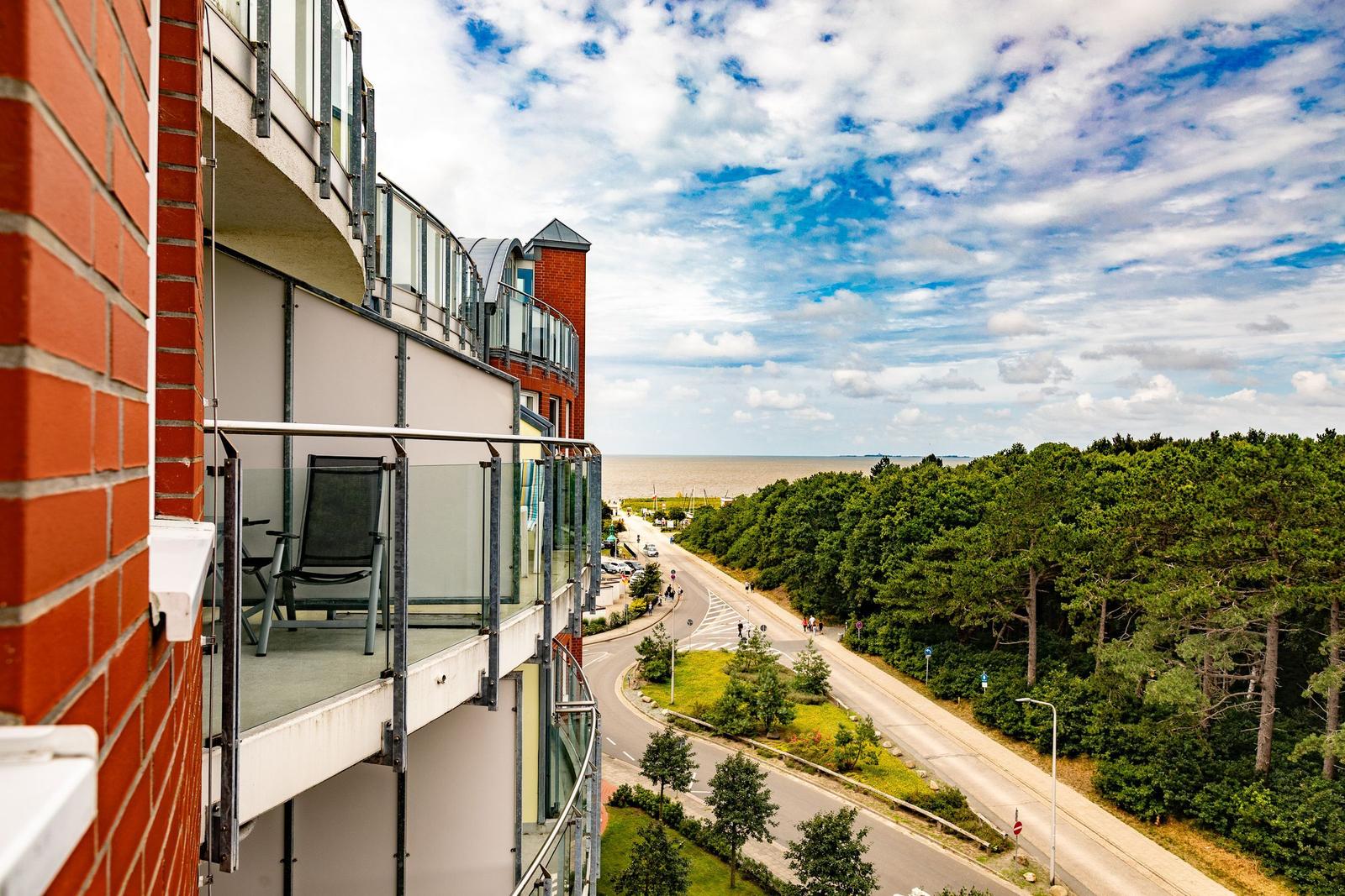 Blick von Balkon auf Straße, Wald und Meer unter blauem Himmel mit weißen Wolken.