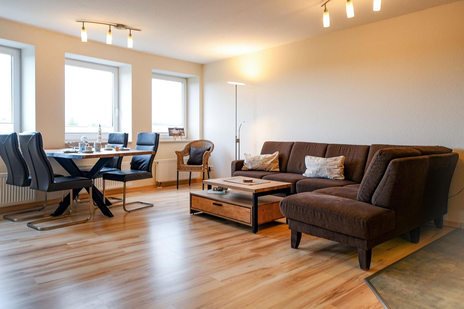 Wohn- und Essbereich mit braunem Sofa, Holztisch und Stühlen, hellen Wänden und Holzboden.
