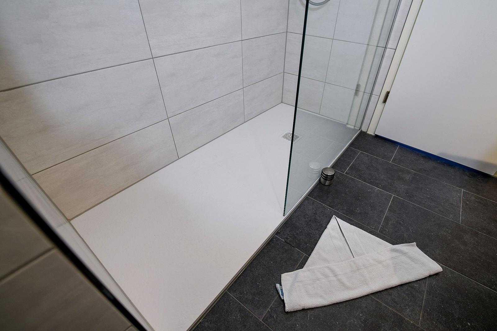 Duschbereich mit Glaswand, weißen Wänden und dunklem Boden. Handtuch liegt auf dem Boden.