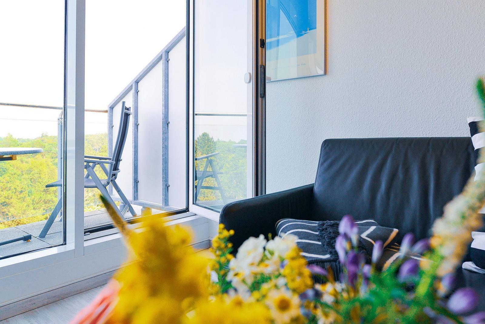 Schwarzes Sofa vor großem Fenster mit Blick auf Balkon und grüne Landschaft.