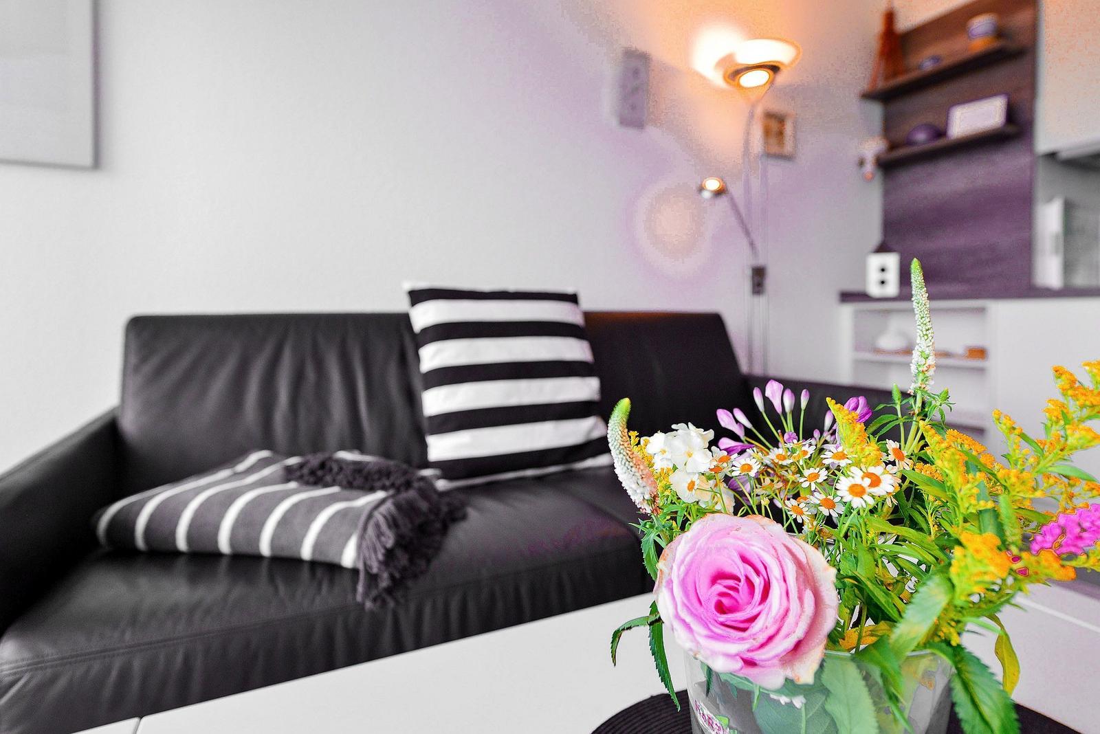 Schwarzes Sofa mit Kissen und Decke, Blumenarrangement auf Tisch, Wandlampe und Regale im Hintergrund.