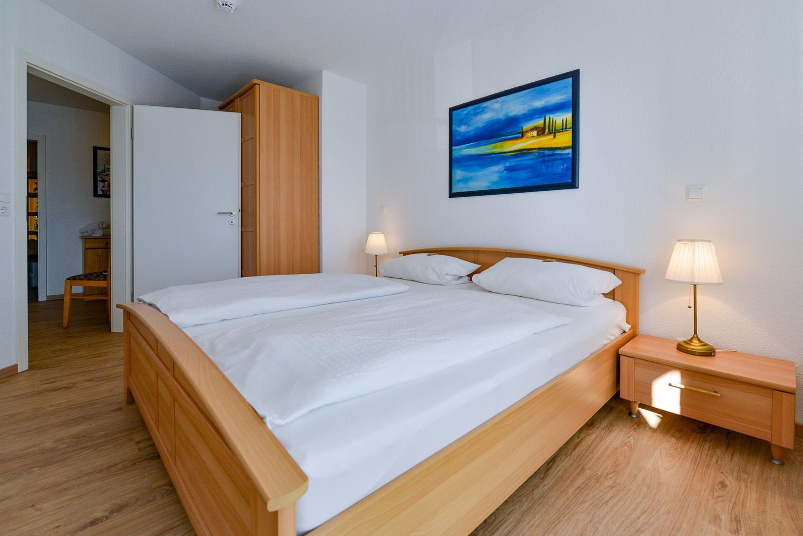 Schlafzimmer mit Holzbett, Nachttisch und Wandbild. Tür führt in angrenzenden Raum.