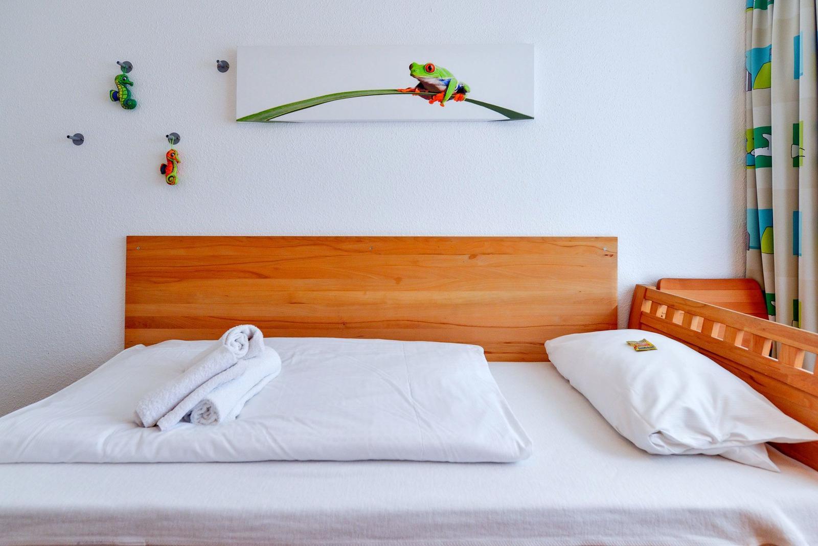Ein Bett mit weißer Bettwäsche und einem Holzgestell. An der Wand hängt ein Bild einer grünen Frosch.