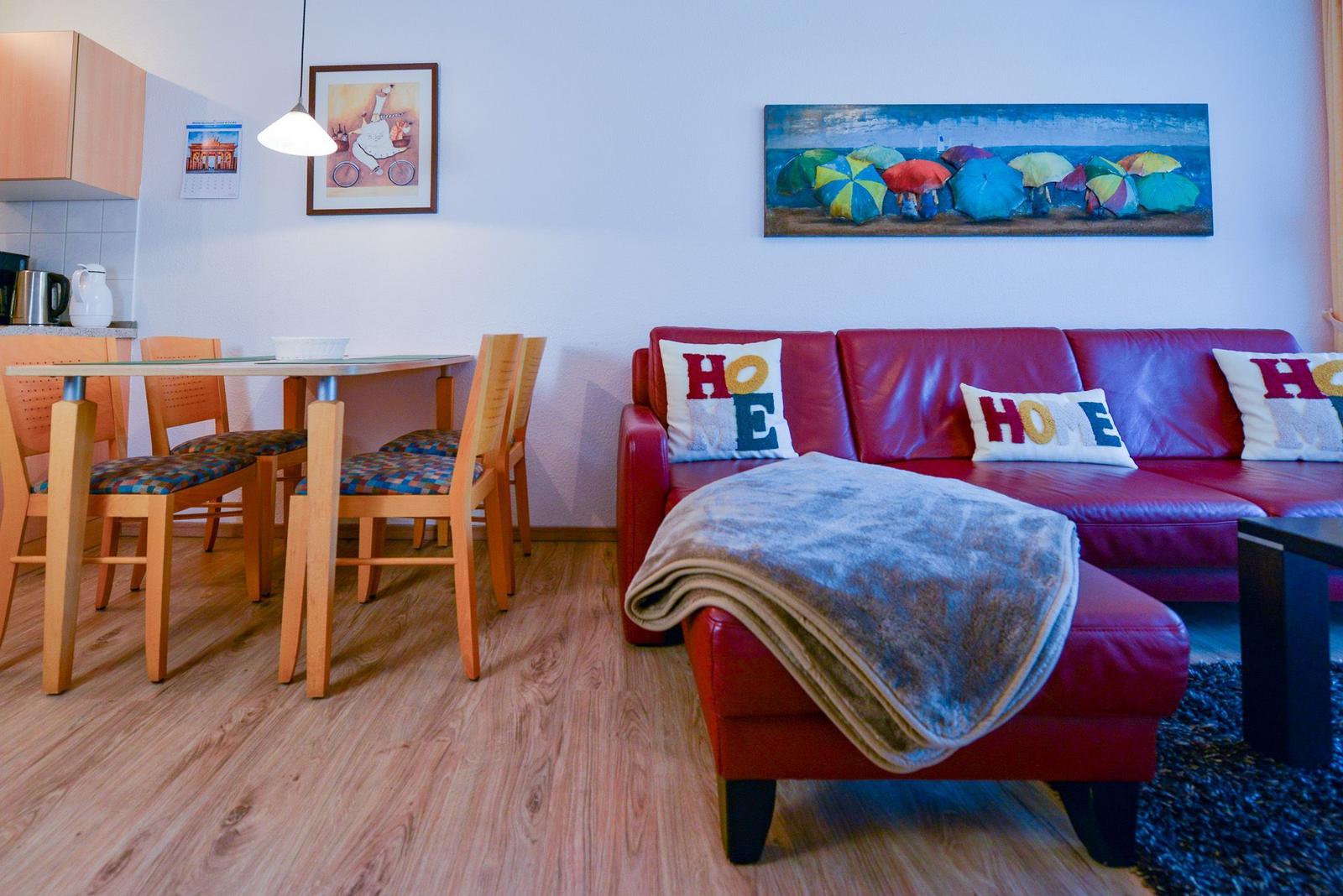 Rotes Sofa mit Kissen und Decke, Essbereich mit Holztisch und Stühlen, Wandbild mit Regenschirmen.