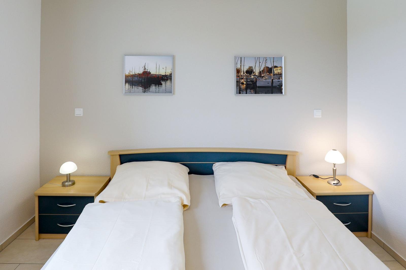 Schlafzimmer mit Doppelbett, zwei Nachttischen und Wandbildern von Hafenbooten.