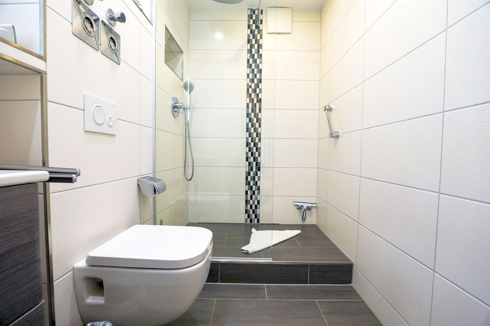 Bad mit Dusche, Toilette und Fliesenboden. Duschwand mit Mosaikstreifen.