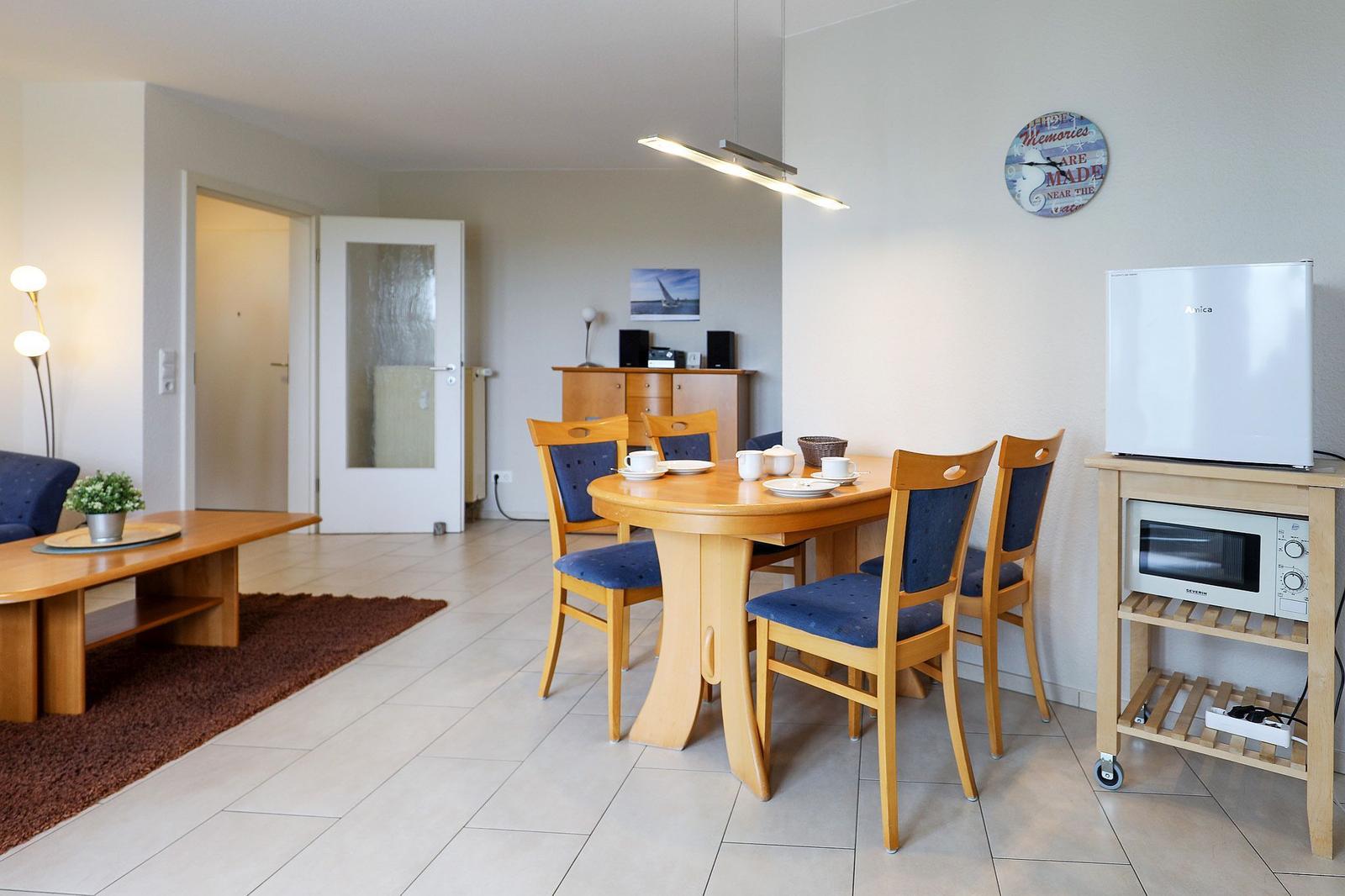 Esszimmer mit runden Tisch, vier Stühlen und Küchenzeile. Im Hintergrund Wohnbereich mit Sofa und Couchtisch.