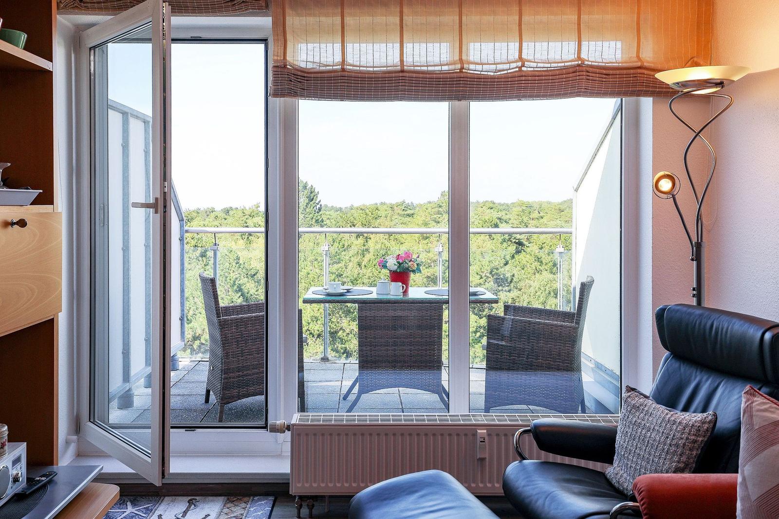 Through the window, a terrace with table and chairs is visible. Outside, there is a green landscape.
