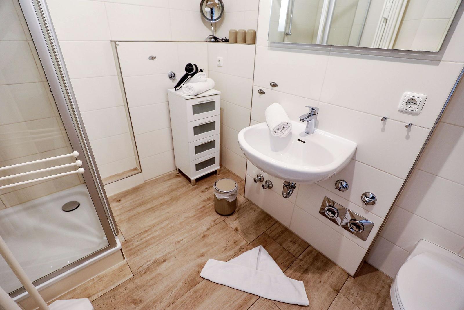 Badezimmer mit Dusche, Waschbecken und Toilette. Holzdielenboden und weiße Fliesen.