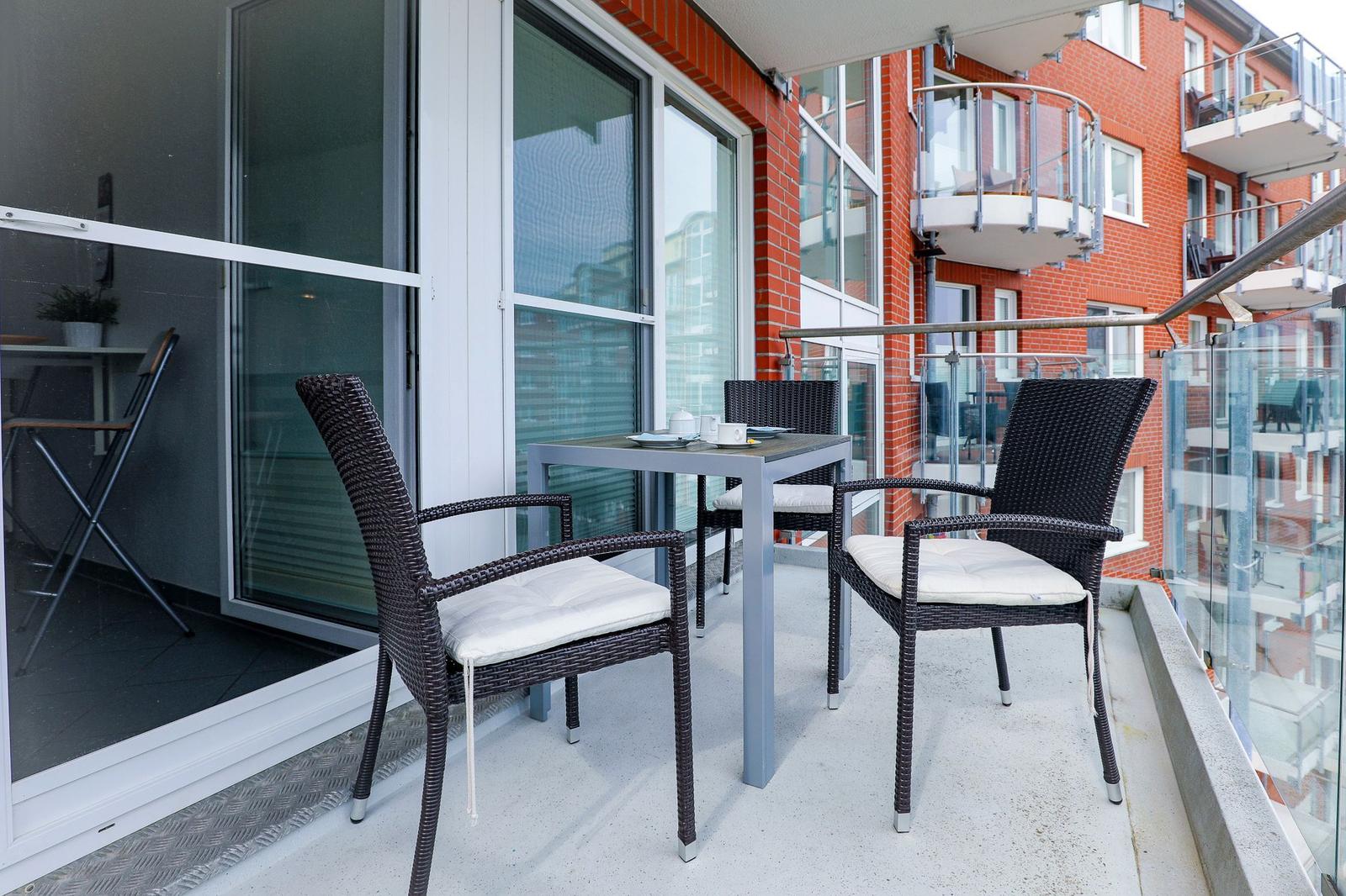 Terrasse mit Tisch und drei Stühlen, Glasgeländer und rotem Backsteinhaus im Hintergrund.