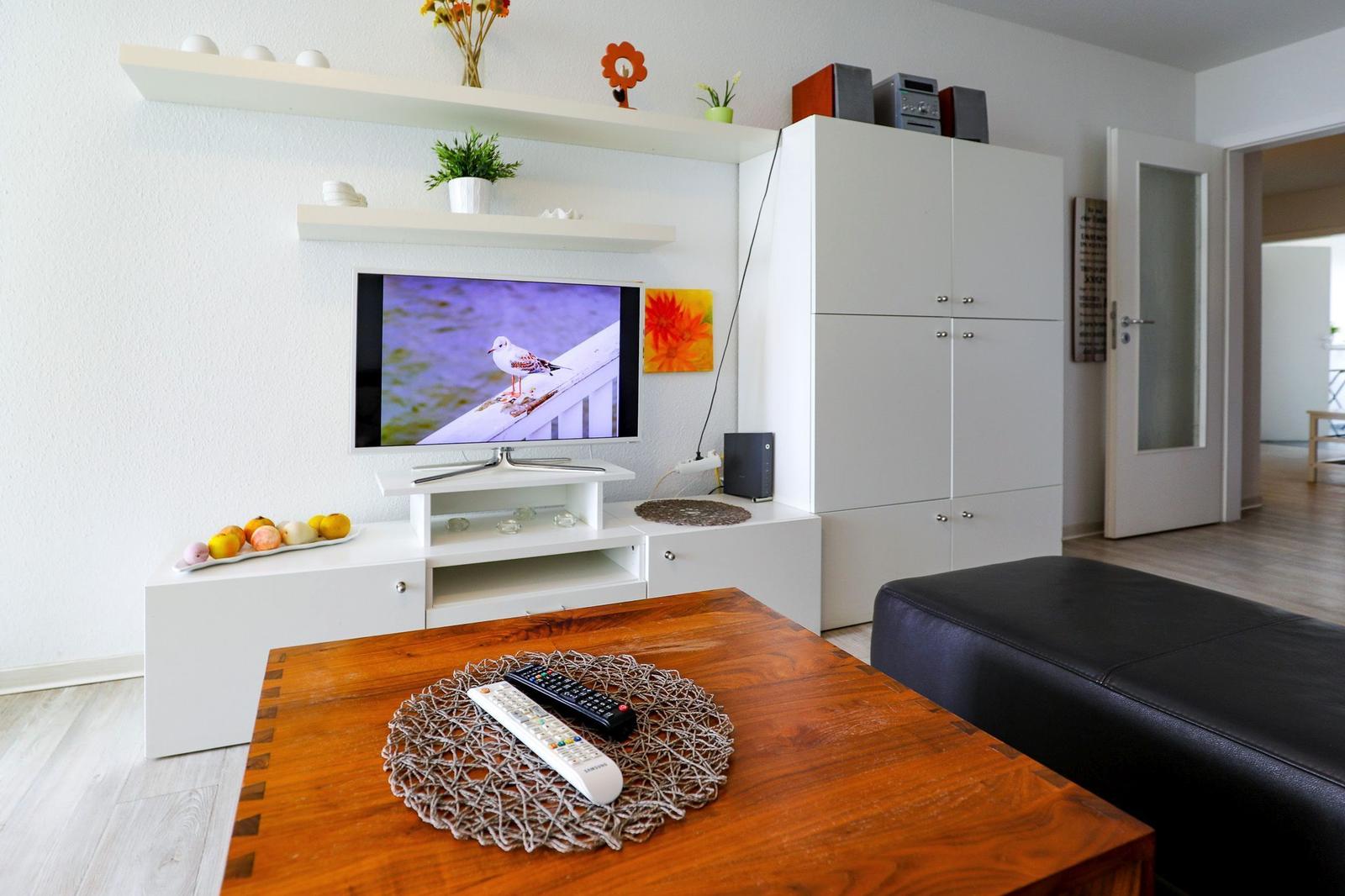 Wohnzimmer mit Fernseher, Holztisch und schwarzer Ledercouch. Weiße Schränke und dekorative Elemente.