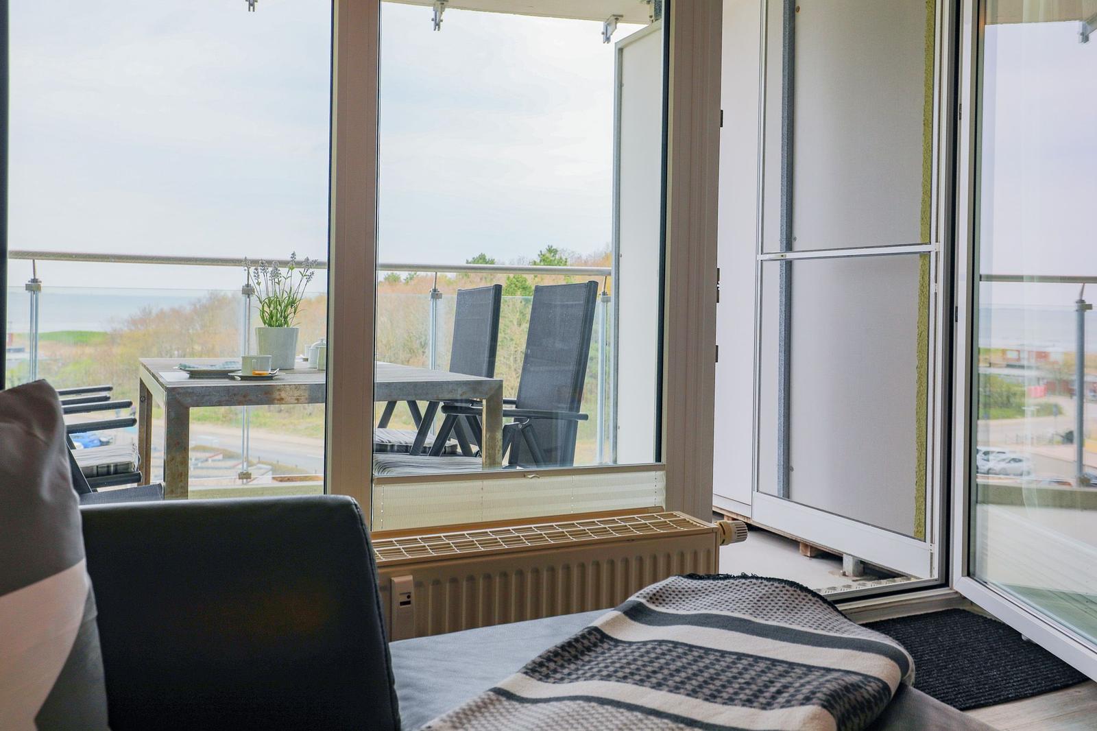 Aus dem Wohnzimmer führt eine Glastür auf eine Terrasse mit Tisch, Stühlen und Meerblick.