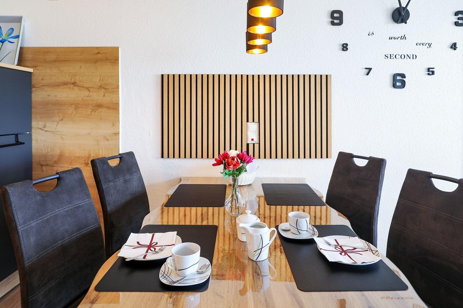 Essbereich mit Holztisch, dunklen Stühlen und Wanduhr mit Zahlen.