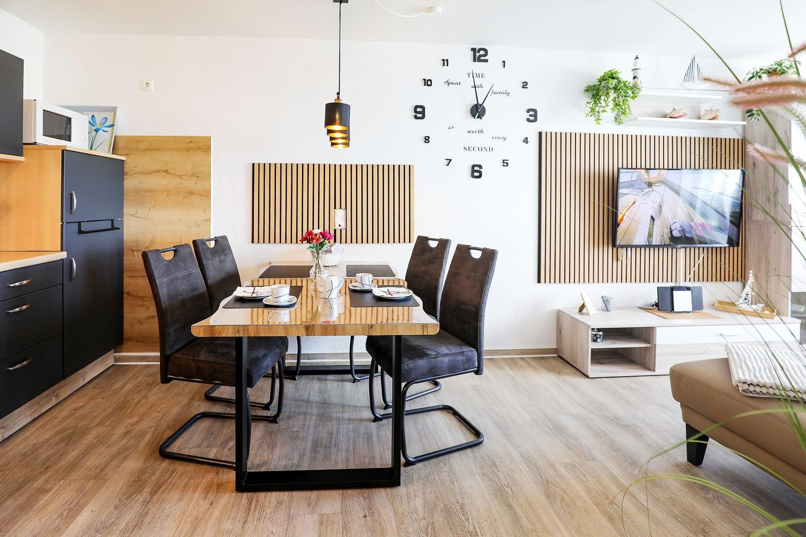 Essbereich mit Tisch und Stühlen, Wanduhr und TV im Wohnbereich.