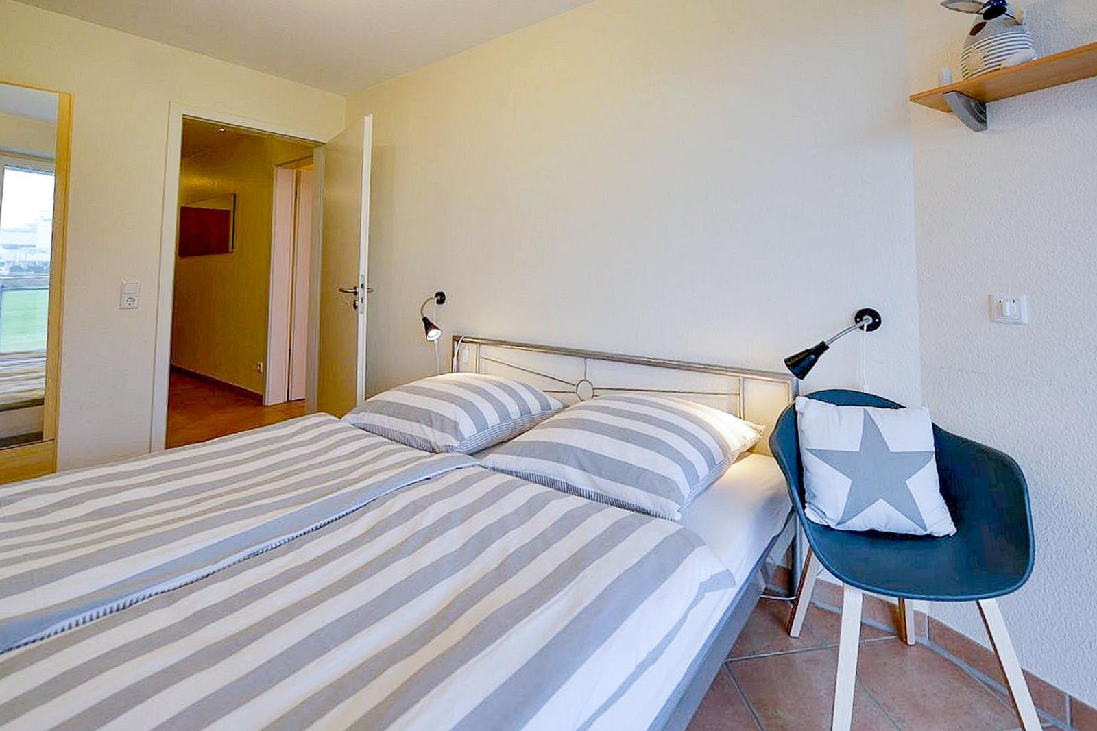 Schlafzimmer mit Bett, Stuhl und Wandlampen. Tür führt in einen Flur.