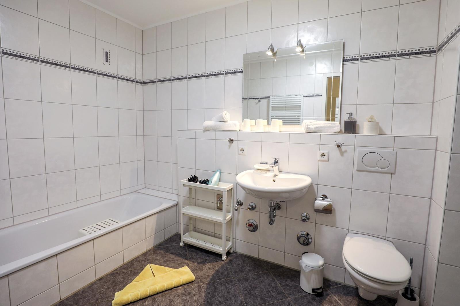 Badezimmer mit Badewanne, Waschbecken und Toilette. Weiße Fliesen und gelber Handtuch.