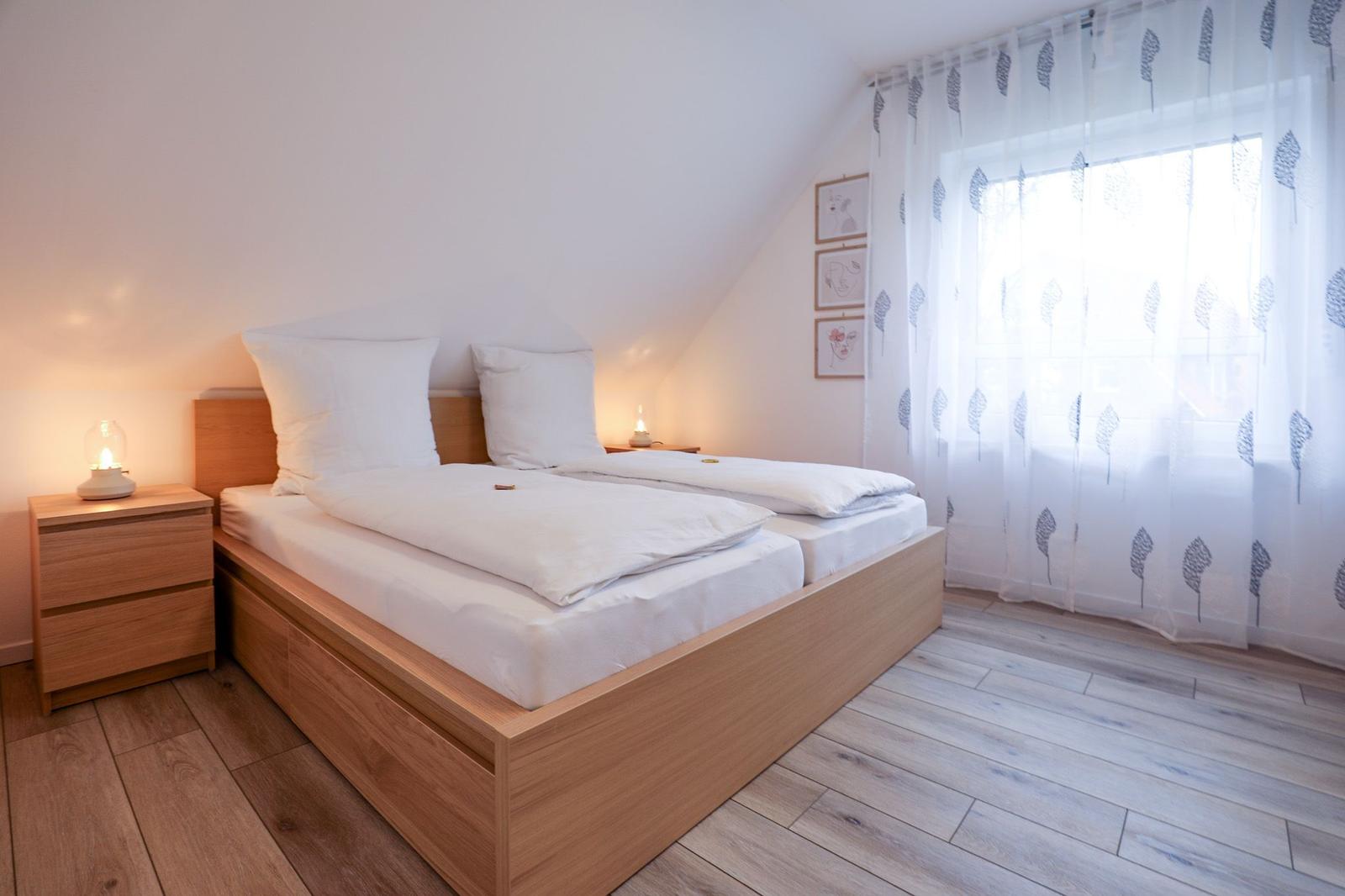 Schlafzimmer mit Holzbett, Nachttisch und Fenster mit Vorhängen.