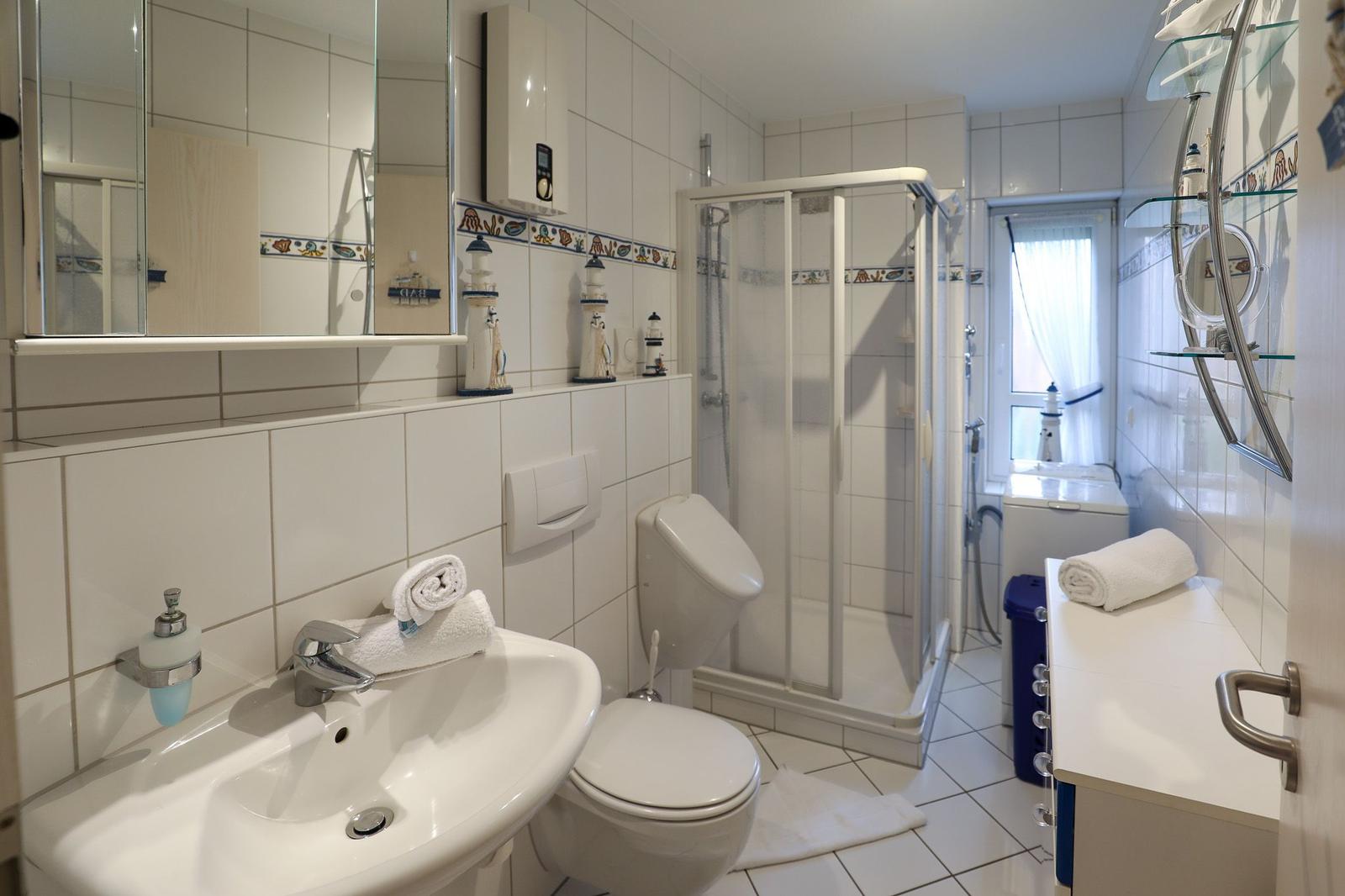 Badezimmer mit Dusche, Waschbecken und Toilette. Weiße Fliesen und Seebad-Dekoration.
