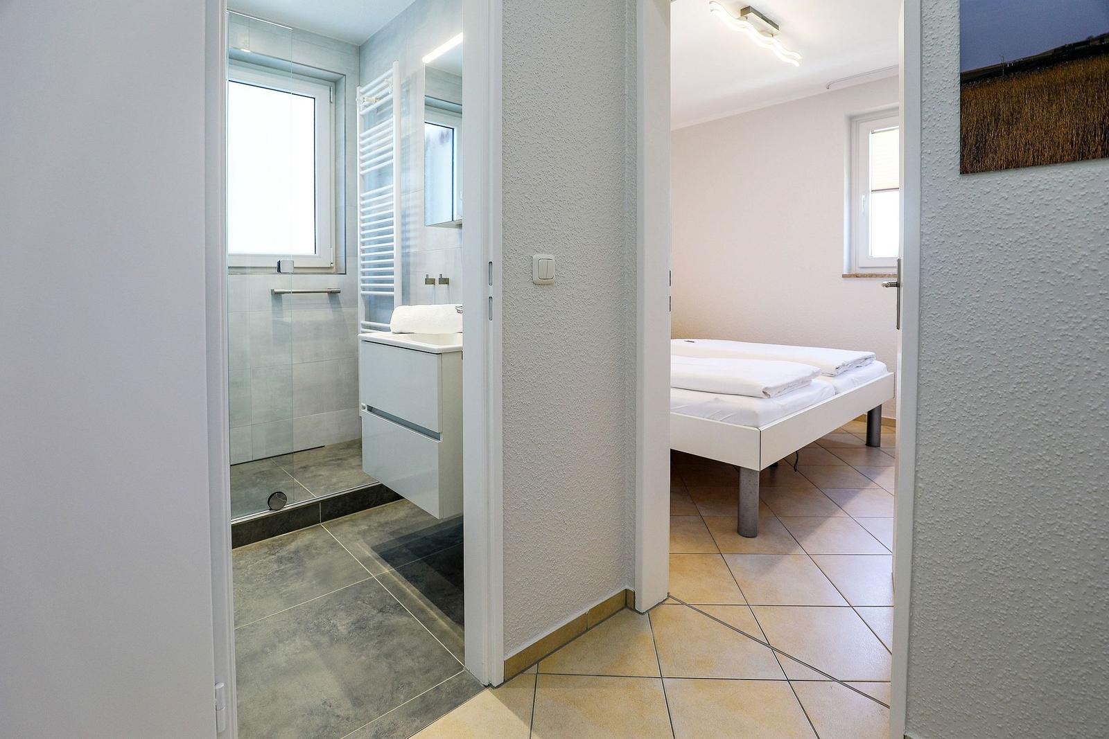 Schlafzimmer mit weißem Bett und Badezimmer mit Dusche und Waschbecken.