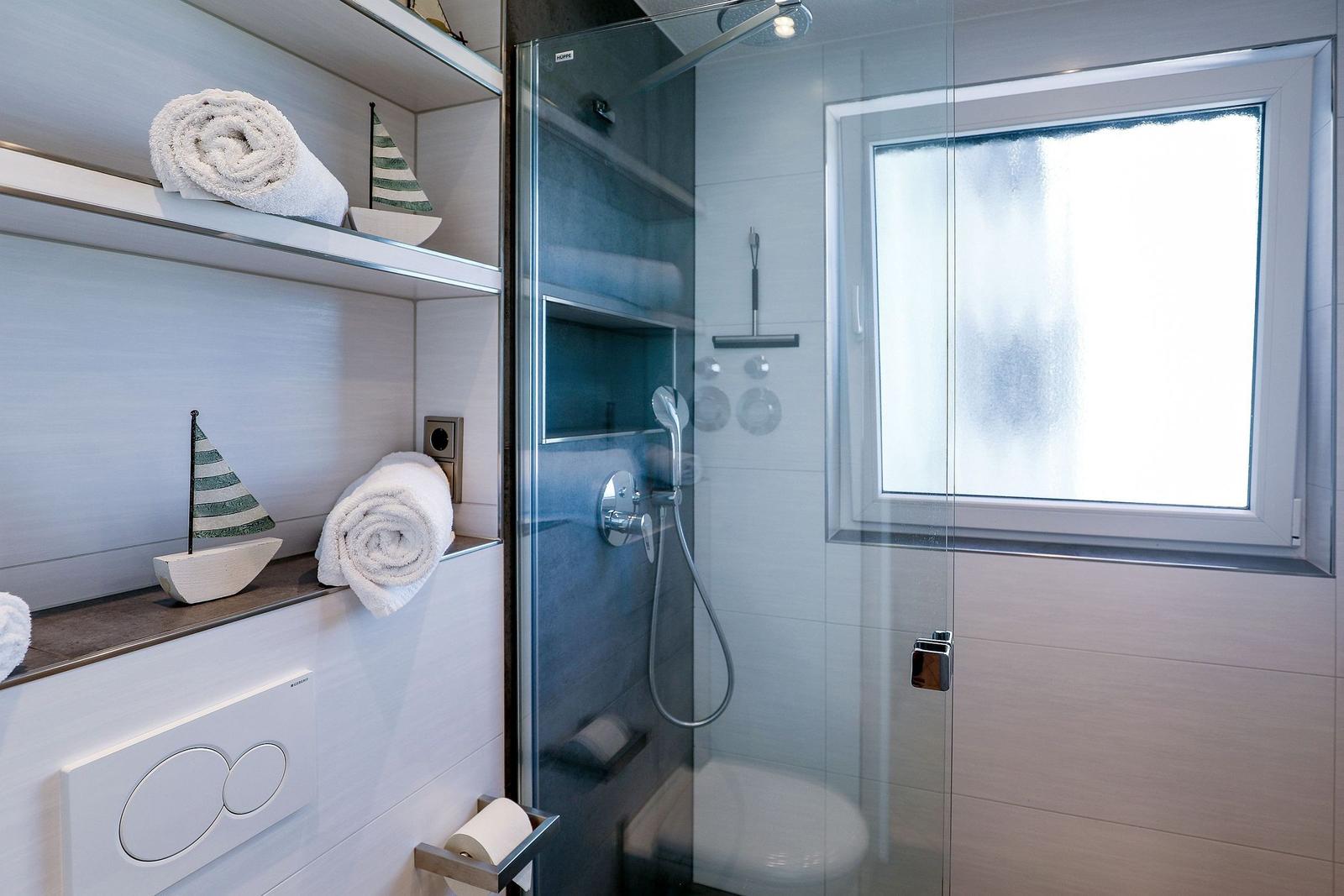 Dusche mit Glaswand, Fenster und Handtuchregal mit Segelboot-Dekoration.