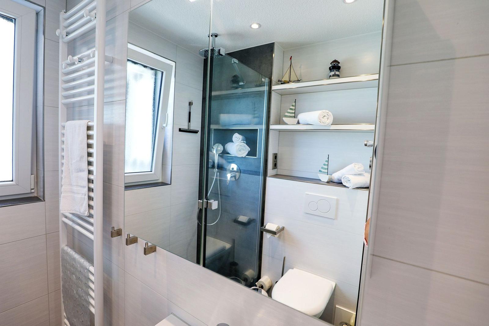 Badezimmer mit Dusche, Spiegelschrank und Heizkörper. Auf Regalen liegen Handtücher und Dekorationsobjekte.