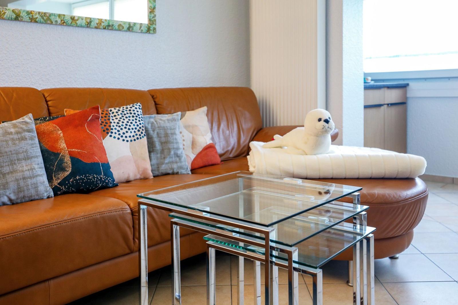 Ein braunes Sofa mit Kissen und einem Stofftier. Davor steht ein Glas-Tisch.
