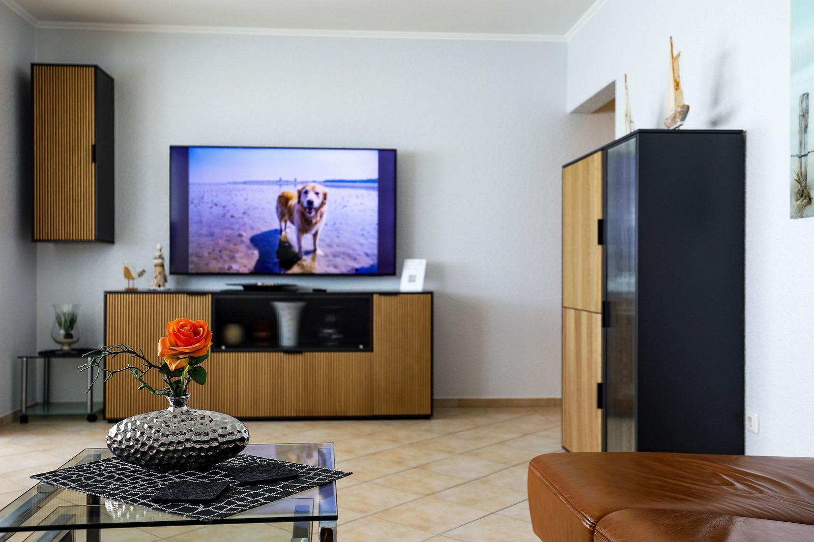 Wohnzimmer mit Fernseher, Couch und Glas-Tisch mit Blume.